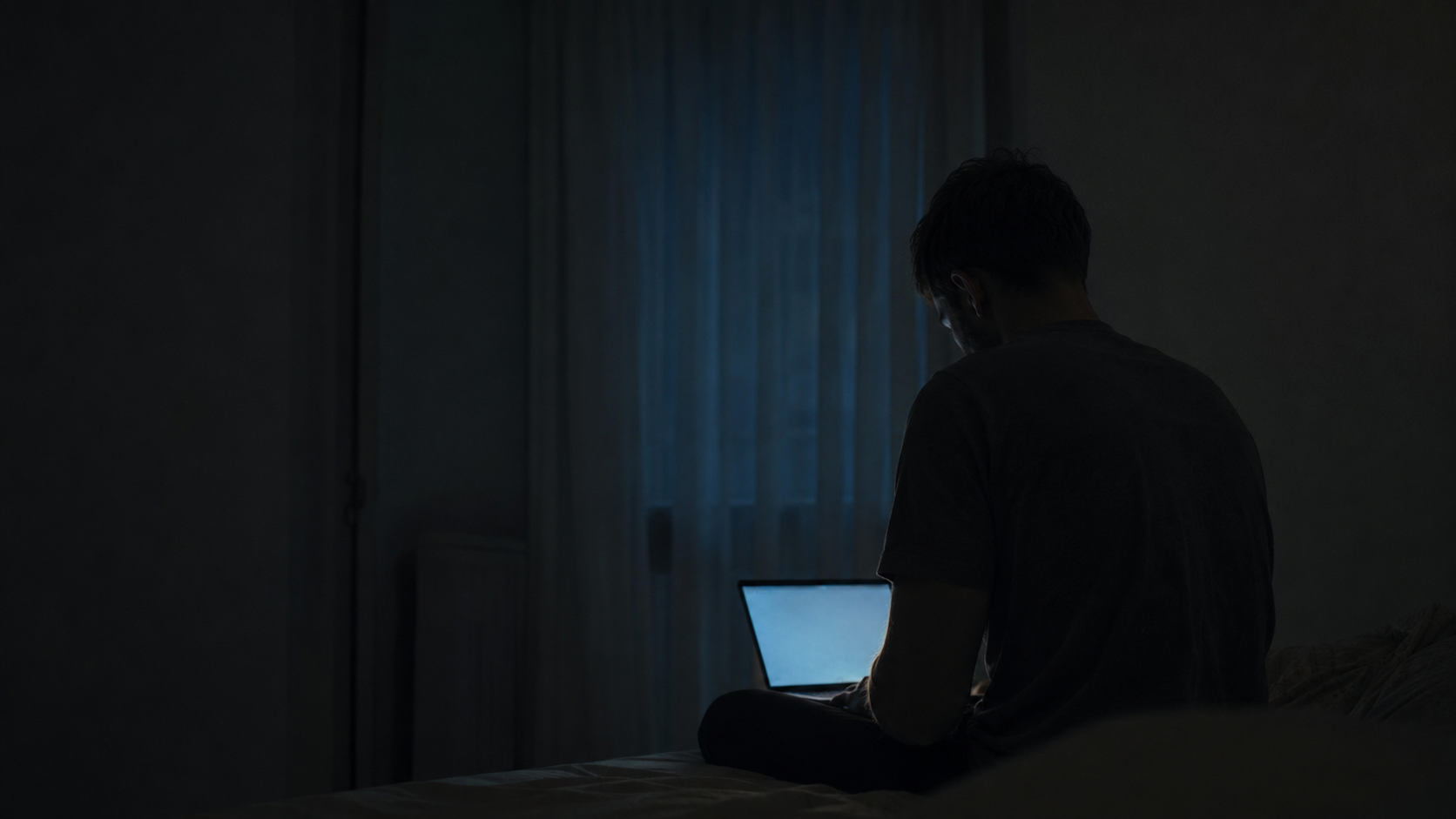 Man sitting alone in a dark room at night using a laptop, illustrating porn addiction, loneliness and isolation